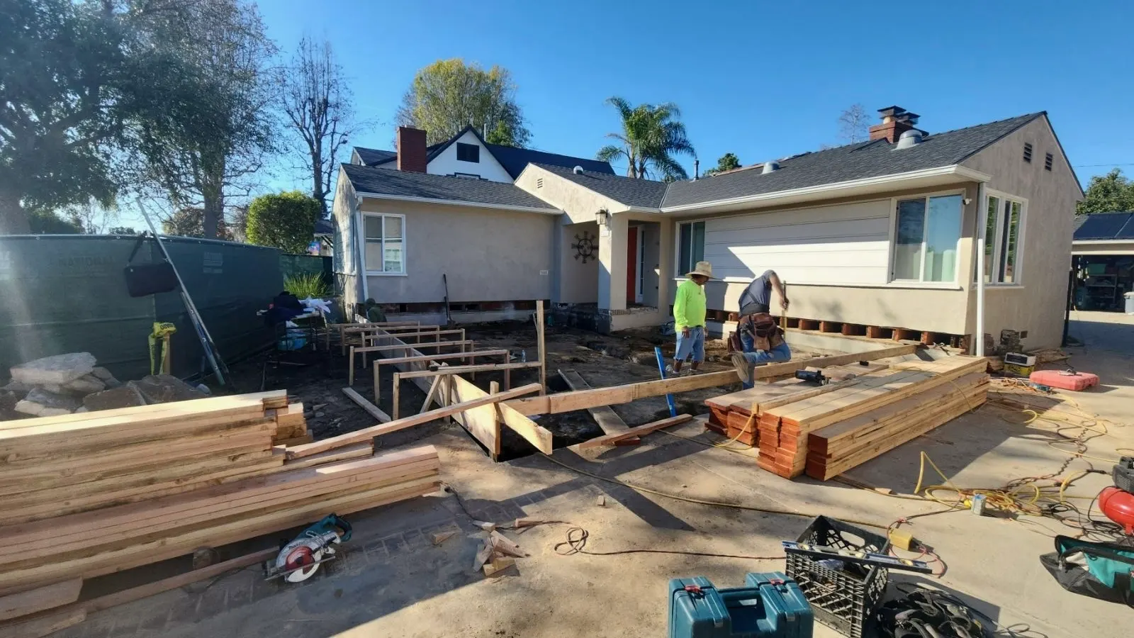 Framing plates set out on a new slab behind a house, with stacks of lumber alongside and two of the crew working