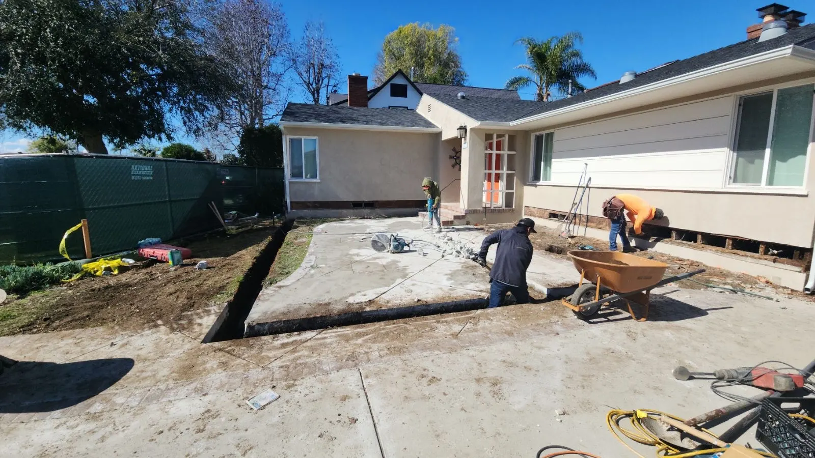 A rear yard with the old concrete patio broken out and a footing trench dug along the back of the house, two of the crew clearing it