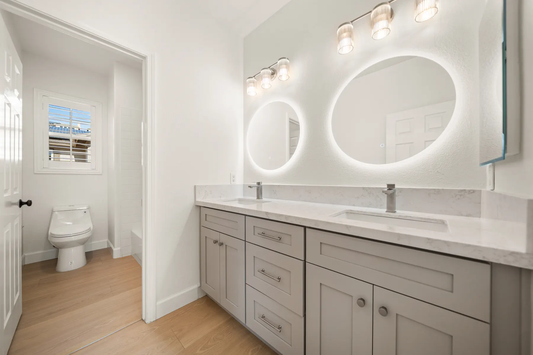 A finished bathroom with a gray double vanity, two round backlit mirrors, and a separate toilet room through an opening