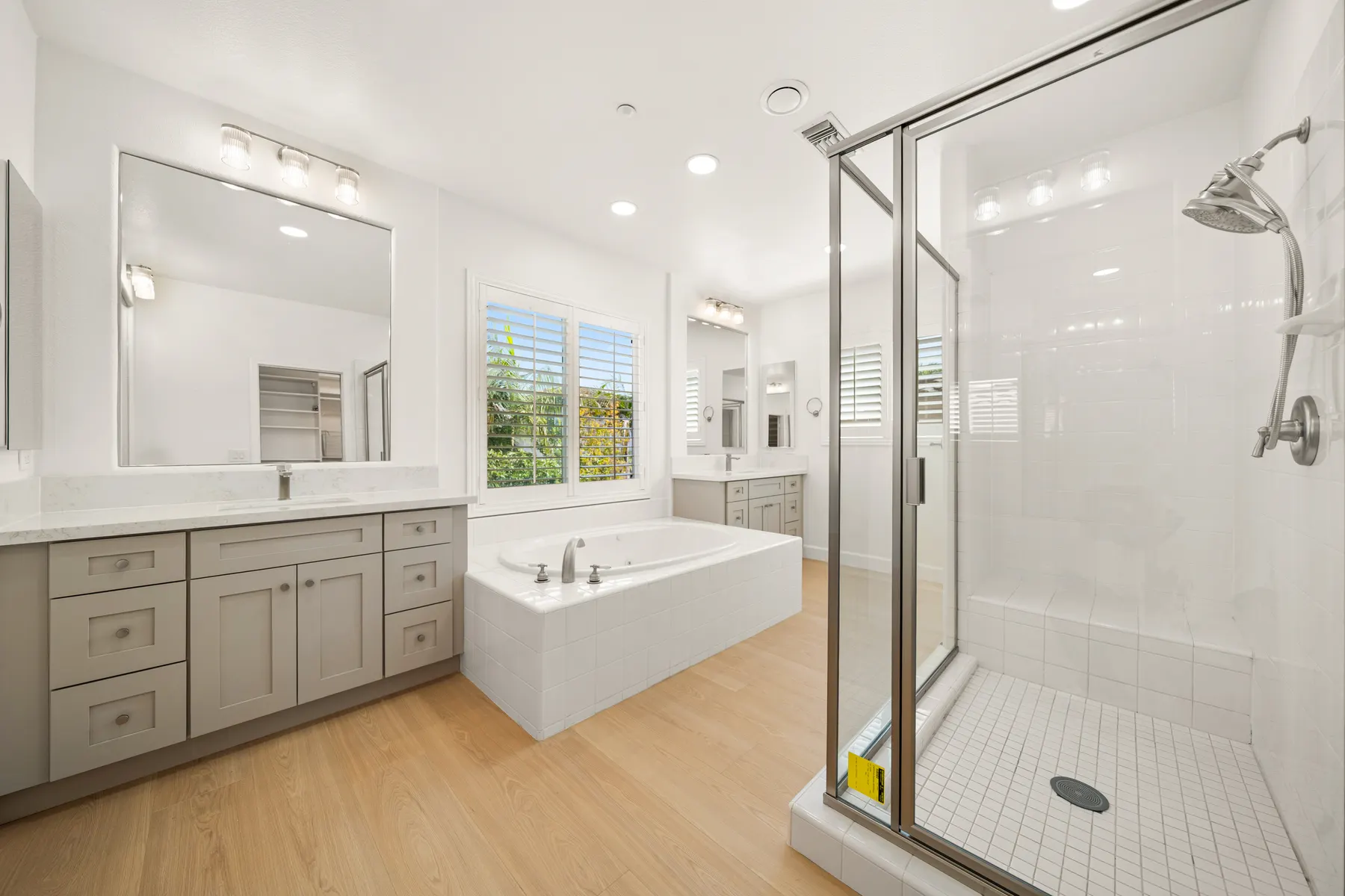 A finished bathroom with a gray shaker vanity under a large mirror, a drop-in tub below a shuttered window, and a framed glass shower in white tile