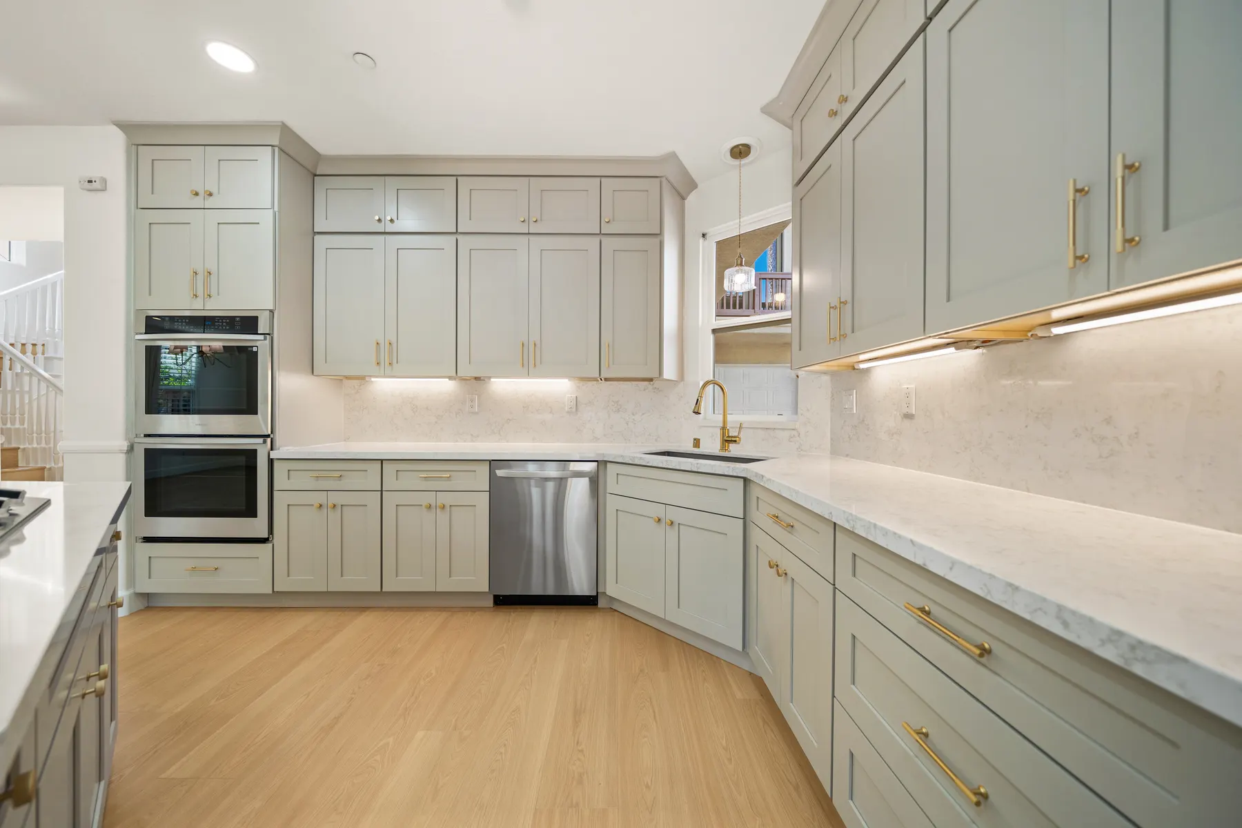 A run of sage-green cabinets with brass handles, double wall ovens, a dishwasher and an undermount sink below marble-veined counters