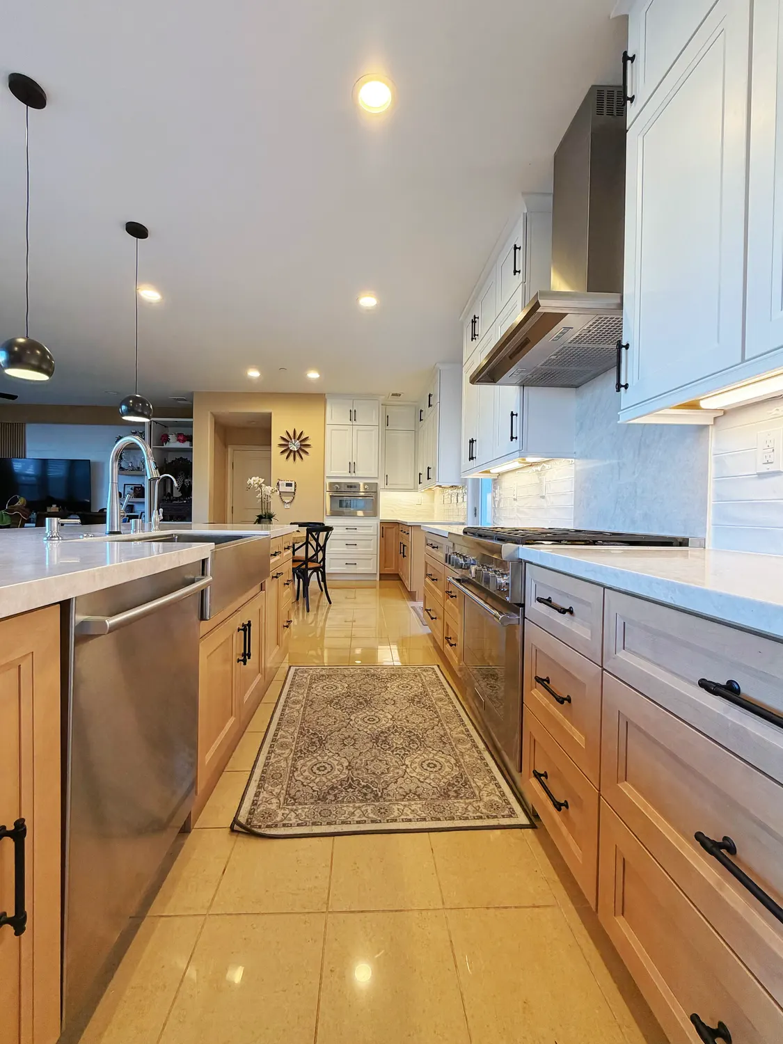 A kitchen with white upper cabinets over natural oak bases, a stainless range under a steel hood, and a patterned runner along a limestone tile floor