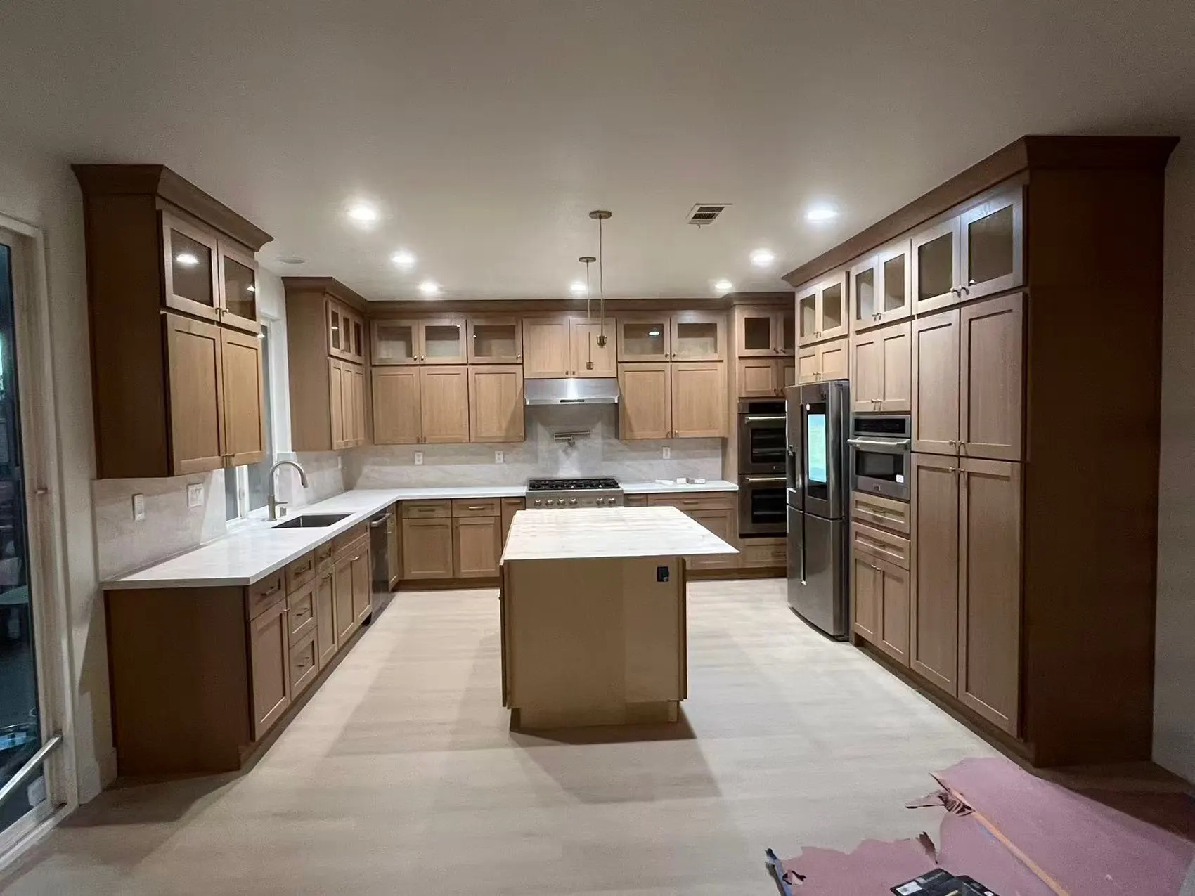 A kitchen in mid-brown raised-panel cabinets with glazed upper doors, white stone counters and a small center island, protective paper still down on the floor