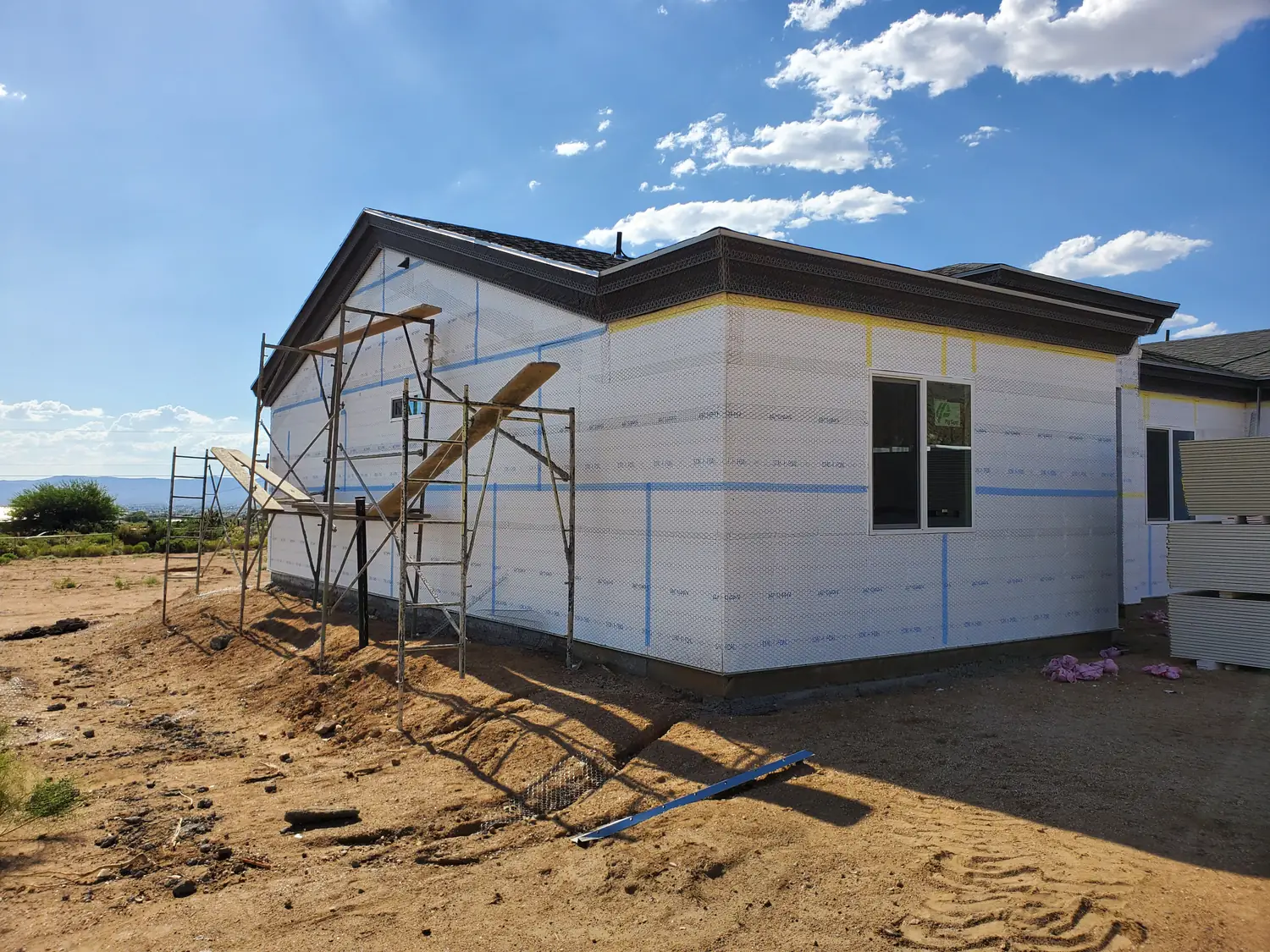 A single-story new build wrapped in housewrap with scaffolding up one gable end, standing on open ground