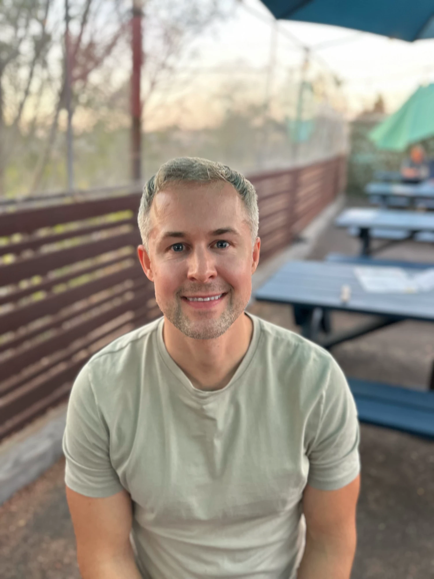 Dane Ziperski in a sage T-shirt on an outdoor patio, picnic tables blurred behind him